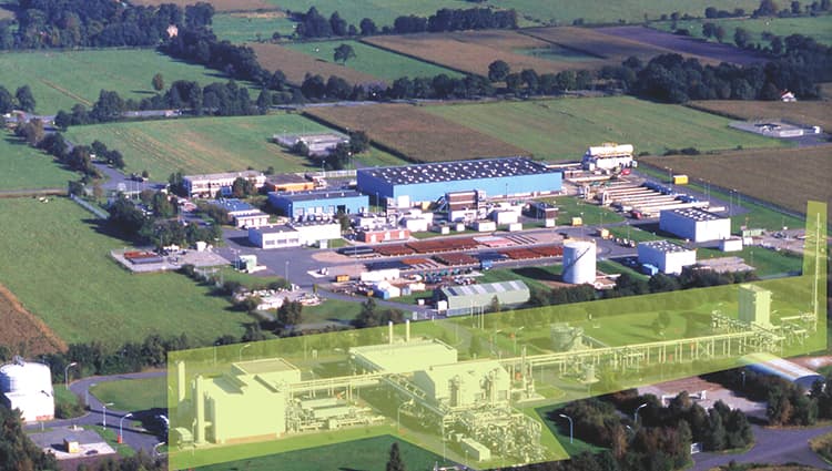 Aerial view of a large industrial plant with multiple buildings and smokestacks, surrounded by green fields.