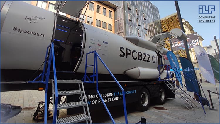 A mobile SpaceBuzz rocket simulator parked outdoors, with stairs leading up to the entrance and various sponsor logos on the side, including ILF Consulting Engineers. The exhibit is set up in an urban area with visitors entering the capsule.